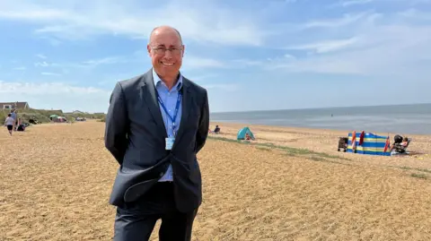 A man in a navy suit, no tie, stands on a golden sandy beach with his arms behind his back. 