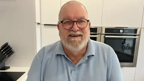 A man in his sixties with a grey beard and black glasses smiles into the camera in his white kitchen.