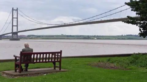 Geograph Humber Bridge viewing area on Hessle Foreshore, with a man sat on a bench on a grassy area next to the river 