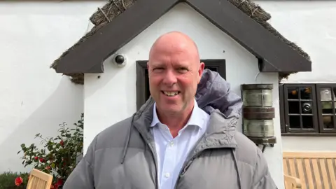 A bald man wearing a white shirt and grey coat stands in front of the entrance to a white building.
