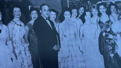 Submitted A black and white photograph from the 1950s of a group of young women in ball gowns smiling and a man in a bow tie and suit looking straight-faced as he holds his hands in front of him.