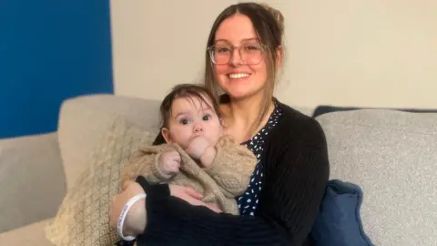 Woman with dark hair, glasses, smiling at the camera and holding her baby