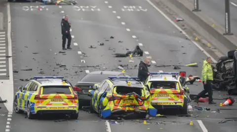 PA Media Three police vehicles, smashed with debris all over three lanes of an A road. A black BMW can be barely seen in front of the middle police car. Three men walk across the road, assessing the scene, in police uniforms.