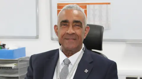 Paul Yates is wearing a dark suit and smiling at the camera while sat behind a desk. 