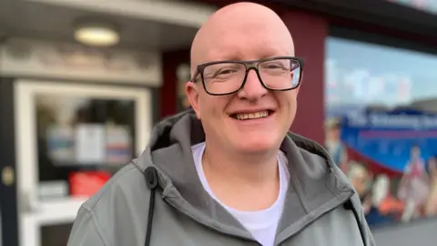 A man with black rimmed glasses is smiling at the camera. He is wearing a grey coat and is standing outside a building.