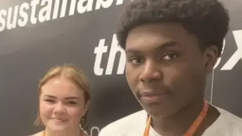 A woman with blond hair, a light brown cardigan and an orange lanyard alongside a man with black hair a white T-shirt and an orange lanyard, standing in front of a black wall with a picture of a person's internal organs underneath a blue outline