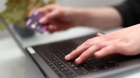 PA Media A person is using a laptop to complete an online purchase. One hand is typing on a keyboard, the other is holding a bank card 