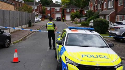 Dawid Wojtowicz/BBC A police officer and police car with police tape cordoning off a street of 1960s style red brick houses