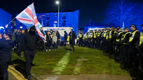 PA Media Police officers form a line in front of a group of protesters with police vans in the background