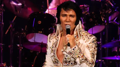 A man with thick black hair and sideburns sings into a microphone. He is on a stage, with a drum kit behind him. He wears an open necked top covered in gemstones