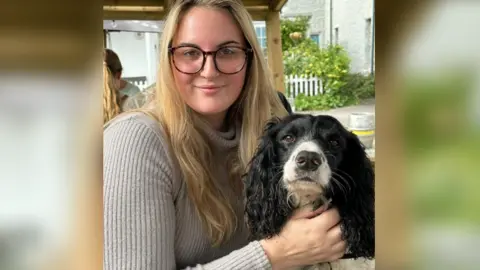 Holly Crouch Holly Crouch, una mujer joven con cabello rubio largo y ondulado y gafas de tortuga, lleva un jersey gris con un cuello alto mientras sostiene a su perro, un spaniel blanco y negro, y sonríe a la cámara.