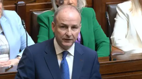 Oireachtas TV A man in a blue suit and blue tie stood speaking in the chamber