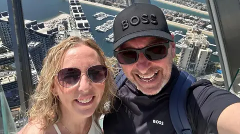 Cynnamon and Tony Church smile from the top of a building overlooking a marina in Dubai. Tony has a black Boss baseball cap on and is wearing a dark Boss T-shirt with a rucksack on his back. Cynnamon, who has curly blonde hair, is wearing a white top. Both are wearing sunglasses.