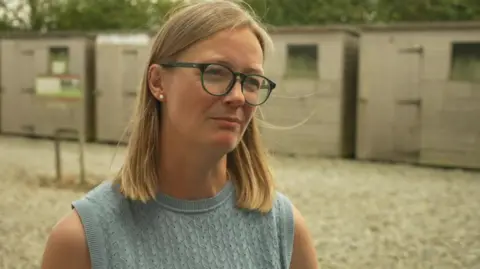 Ele Brown is wearing a a sleeveless pale blue top. She has shoulder length blonde hair and is standing outside with several sheds in the background.