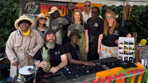 Members of the Windrush society sit on a stall selling T shirts, smiling and holding various items including fans and a pineapple. Many are wearing Windrush society t-shirts.