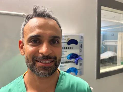 Dr Rastogi is standing in a hospital ward, he is wearing a green surgical smock and is looking at the camera. He has a dark beard with flecks of grey and black/grey hair. He is in his early forties.