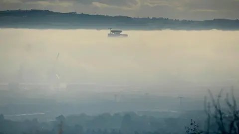 Pacemaker A still, wide image showing a thick layer of fog covering most of Belfast. Sitting above the cloud of fog is the top of the yellow Harland and Wolff cranes.