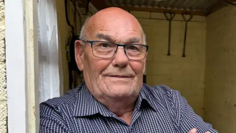 A close up of Terrence Bridges looking straight down the lens. He is leaning against a door frame and a small, brick walled building can be seen behind him. He is wearing a blue checked shirt, which is open at the collar, and black rimmed rectangular glasses. He is bald on top of his head, but some short grey hair can be seen on the sides of his head. 