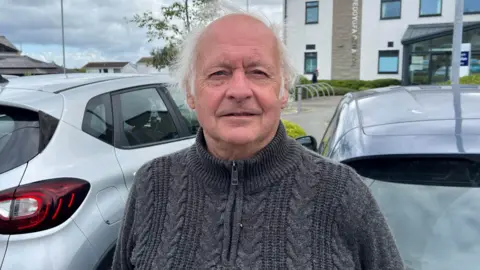 A man with grey hair, stood outside in a car park. He is looking directly at the camera. He is wearing a dark grey knitted quarter zip jumper, with a high neck.