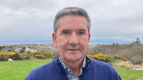 Daniel O'Donnell is wearing a blue fleece, a white and blue shirt and is standing behind a backdrop of large green fields and trees. A number of houses are visible in the background.
