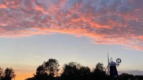 Sue P/BBC Pink clouds above trees and a windmill.