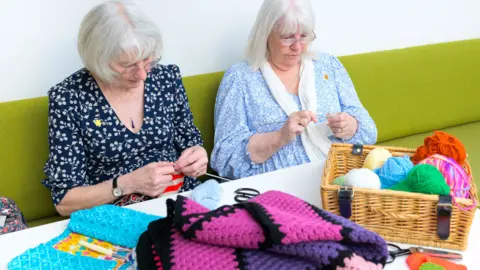 Bristol and Weston Hospitals Charity Two elderly women are sitting on a long lime-green bench and knitting. Both are looking very concentrated through their glasses. There is a table in front of them with a small wicker basket with several balls of different-coloured wool, and a crocheted blanket in pink and purple.