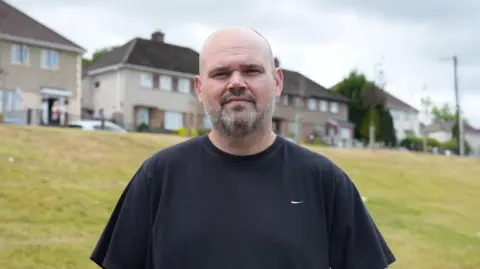 A man with a short trimmed beard is wearing a black tshirt. He's standing at the foot of a grassy bank and in the background are semi-detached houses.