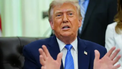 Donald Trump gestures with his hands as he speaks to reporters from the Oval Office while wearing a navy suit with a US flag badge on its lapel, white shirt and blue tie 