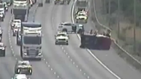 A still taken from a grainy traffic camera. It shows a dark van on its side with people around it. Police cars have blocked it in and traffic is passing to the left.
