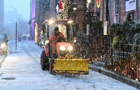 Reuters A man wearing a hi-vis taking driving orange and yellow snow plow with lights on along a snow covered pavement.