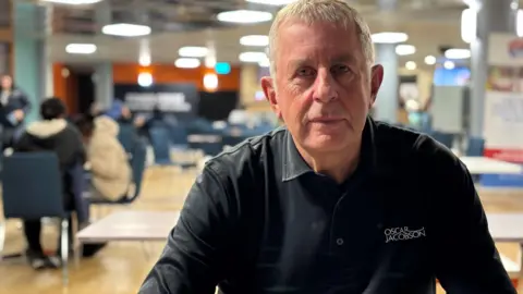 A man is sitting in a large cafe area - the background is blurred but there are tables and about six people sitting on different tables. He is looking directly at the camera with a serious expression. He has short grey hair and is wearing a black shirt, the top button open. 