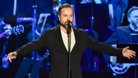 Robin Little/Redferns/Getty Alfie Boe - a man with short brown hair and a beard - sings into a microphone while extending his arms wide to either side in front of band lit blue by stage lights. He is wearing a smart, black jacket over a white T-shirt.