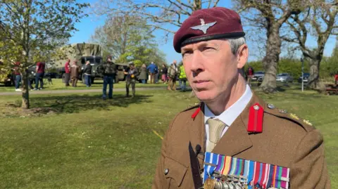 Harry Parkhill/BBC A military man is wearing a burgundy beret with a silver logo on it and a brown coat with a yellow tie and white shirt. He has several medals displayed on the brown coat. Behind him a field can be seen with trees with people dotted about and a green military truck in the distance.