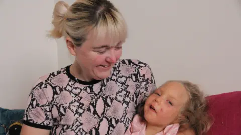 A woman in a pink alligator skin print t-shirt, with bleach blonde hair smiles down at her daughter, who has honey blonde hair, and looks at the camera. 