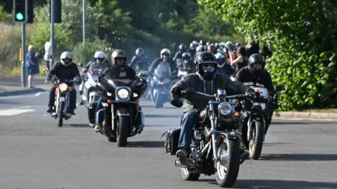 A large convoy of motorcyclists driving down the road, with puffs of smoke coming from their exhausts. 