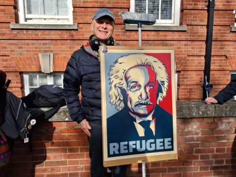LDRS A man wearing a blue padded jacket and baseball cap holding a placard with a depiction of Albert Einstein on it. It has the word "refugee" at the bottom.