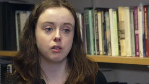 The head and shoulders of a young woman with shoulder-length brown hair,   pictured in front of a bookshelf