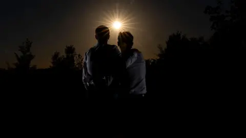 Jerry and Jamie are in silhouette as the moon clouds the sun. They each have an arm around each other as they gaze towards the eclipse. 