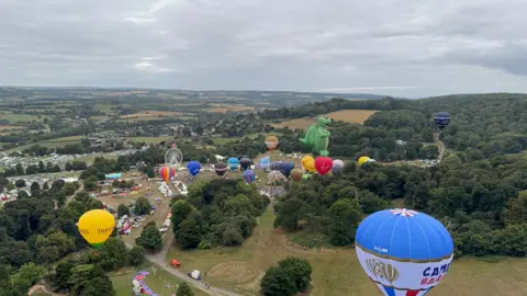 Several colourful hot air balloons taking off from a field in Bristol on an overcast day. They are surrounded by fields and trees.