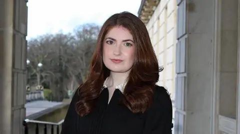 Connie Egan, a woman with long, brown, wavy hair, looks directly at the camera while standing outside Parliament Buildings at Stormont. She is wearing black dress with a white collar