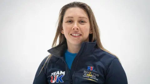 PA Media A woman with long dark blonde hair is wearing a navy blue fleece over a turquoise top. Her fleece is labelled with Team UK and Invictus Games badges.