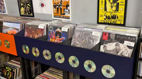 BBC/Eve Connor Album-sized vinyl records stacked in a dark blue rack with four sections, next to a similar orange rack which only partially seen. The blue rack has seven silver CDs stuck on the front in a row for decoration. The album covers show names such as Steely Dan and Tori Amos. Behind the racks concert posters are on the wall in black or white frames.