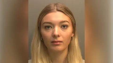 Lincolnshire Police A police custody photo of Madeleine Lonsdale. She has long blonde hair and is wearing a grey t-shirt and small gold hoop earrings. The wall behind her is grey.