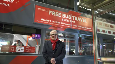 Richard Parker, Mayor of the West Midlands, promoting a pre-Christmas offer of free evening buses. He is standing next to a ticket office window and there is a red and white sign advertising the scheme above him. He has grey hair and glasses and is wearing a black jacket and red scarf.