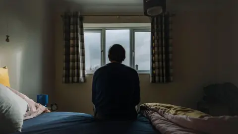 An anonymous man sits on bed and looks out window in a depressing scene 