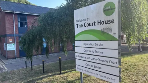 Coroner's court in Ampthill: A white noticeboard which includes the name "Bedfordshire and Luton Coroners Court", written in black letters, standing on green grass outside a red-bricked building.