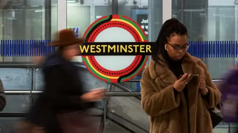 GG Archard A Westminster tube station roundel in the Pan-African colours, with a lady in a faux fur coat standing in front of the roundel looking at her phone and another person out of focus walking along the platform. 