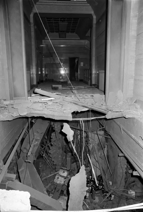 PA Media A black and white image showing a street-level ceiling blown away and a cavity into an underground room. Rafters and wiring hang down and rubble and bottles are strewn across the floor.