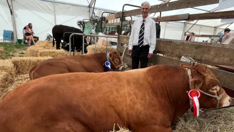 Ann Atkins Colin Atkins standing next to two large, light brown bulls. They are perched on some hay inside a white marquee. Mr Atkins is wearing a white shirt and burgundy striped tie. He is smiling.