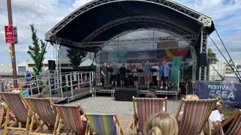 Festival of the Sea A large stage with a group of people on. There are colourful deckchairs in front of the stage. 
